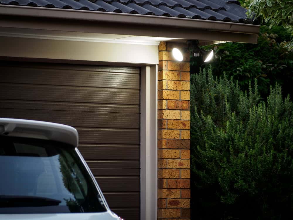 A garage with a brown door illuminated by two bright outdoor lights positioned above it on a brick column. A white vehicle is partially visible in the foreground, and lush green shrubs are seen to the right of the garage.