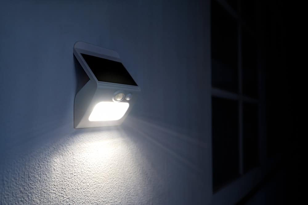 A solar-powered motion sensor light illuminated at night, attached to an exterior wall next to a window, casting a bright glow. The wall is painted white, reflecting light around the fixture.