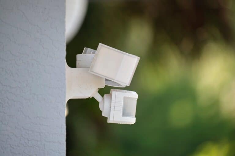 A white motion-sensor floodlight is mounted on the corner of a building with a textured concrete surface. The background is blurred with green foliage, suggesting an outdoor setting.