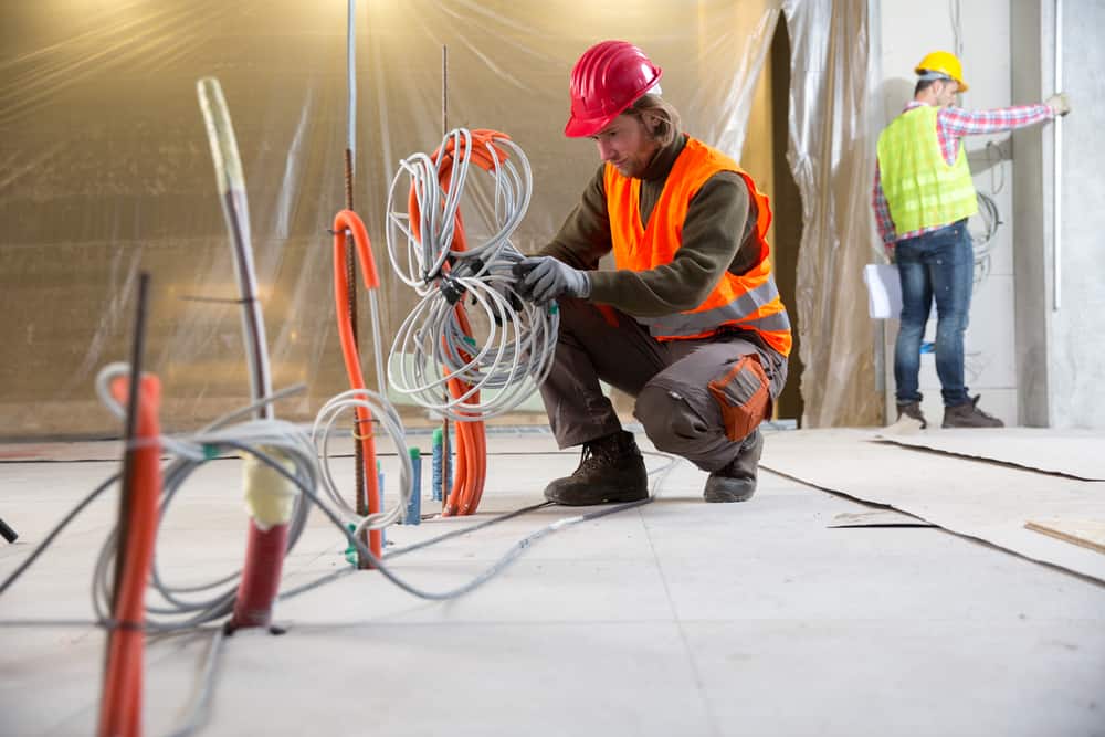 A worker in an orange safety vest and red hard hat crouches on the ground, handling electrical wires at a construction site. Another worker wearing a yellow hard hat and reflective vest stands in the background holding papers, near partially installed walls, showcasing the expertise of electrical contractors Long Island.