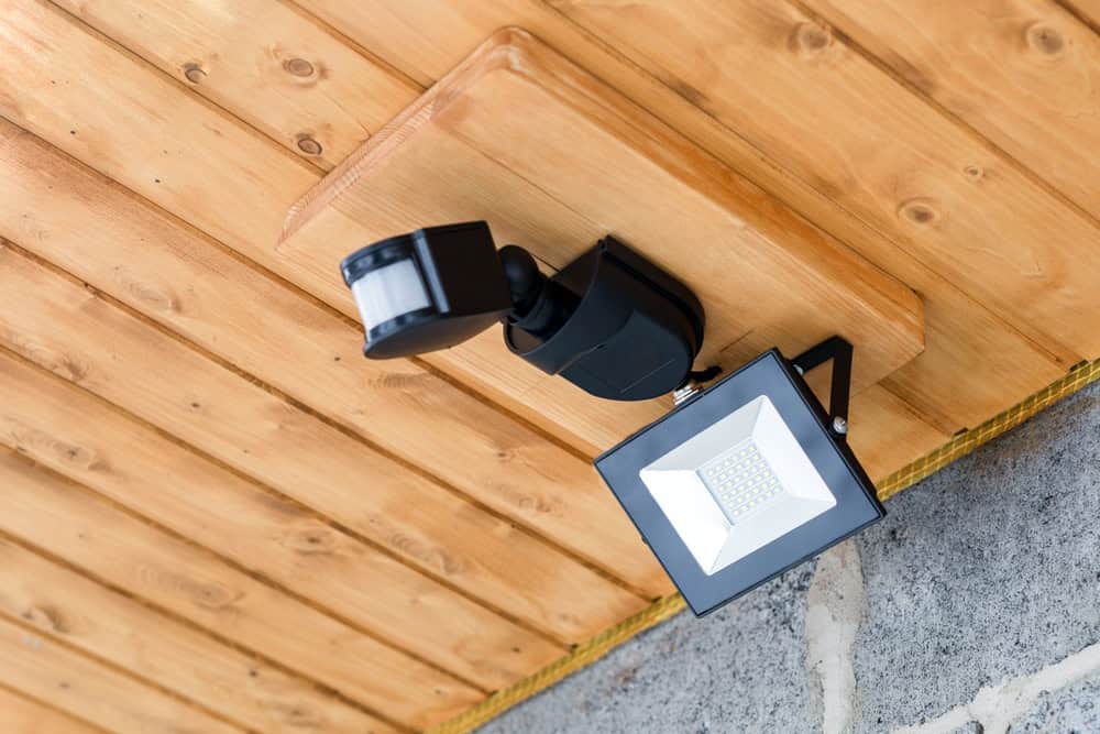 A motion sensor security light mounted on a wooden ceiling. The light is positioned on the right, while the sensor extends from the left, both attached to a square base. The ceiling features a natural wood finish.