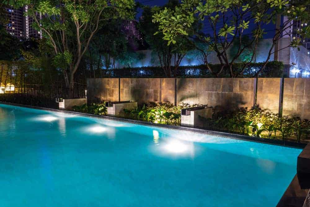 A serene night view of a swimming pool surrounded by trees and plants. The pool is illuminated by underwater lights, and buildings are visible in the background, softly glowing under night lights.