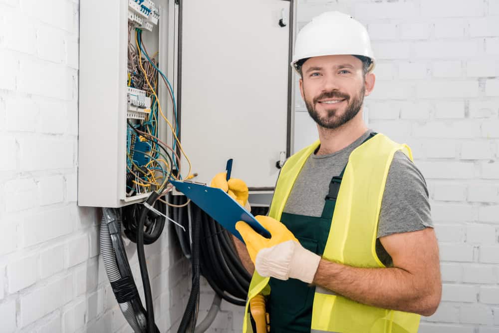 A man wearing a white hard hat, yellow safety vest, and gloves is working on electrical wiring in a control panel. He is holding a clipboard and smiling, standing against a white brick wall.