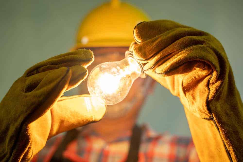 A person wearing a yellow hard hat and protective gloves holds a glowing light bulb. The focus is on the bulb, with the individual in the background slightly blurred. The person is wearing a checked shirt.