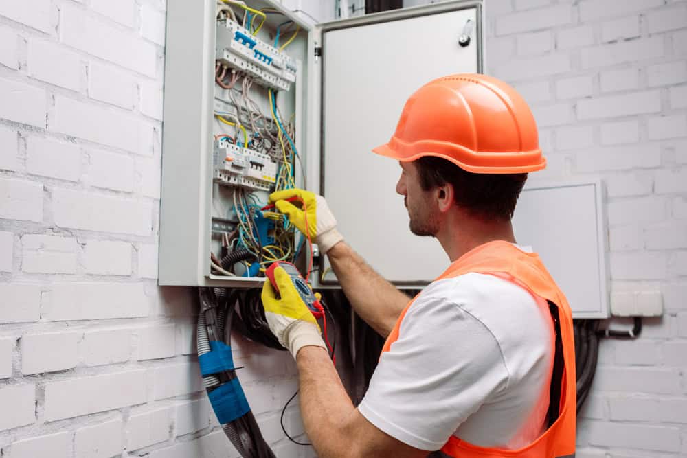 A worker wearing an orange hard hat and safety vest uses a tool to inspect or repair a complex wiring system inside an open electrical panel mounted on a white brick wall.