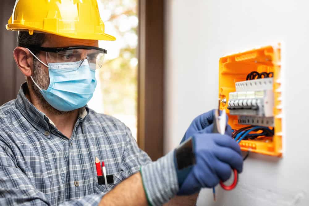 Electrician wearing a hard hat, safety glasses, mask, and gloves, using tools to work on a circuit breaker box mounted on a wall.