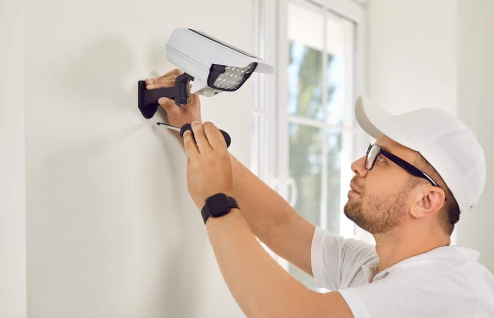 A man wearing glasses, a white cap, and a smartwatch installs a security camera on a white wall. He's using a screwdriver, and a window is visible in the background.