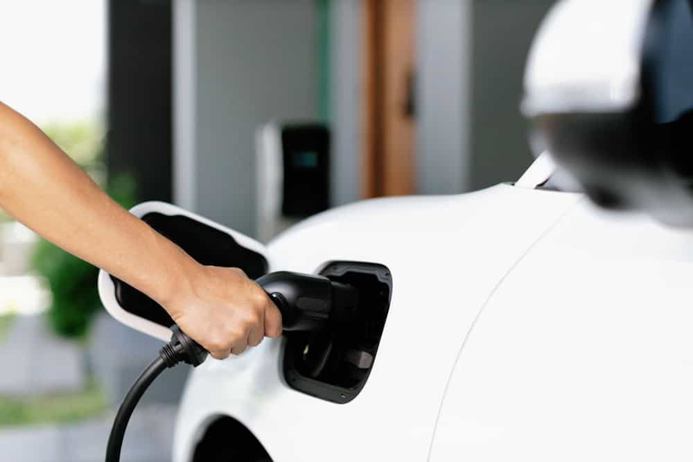 A person plugs a charging cable into an electric car. The vehicle is white, and the focus is on the hand holding the cable and the car's charging port. The background is blurred, suggesting a residential or private setting.