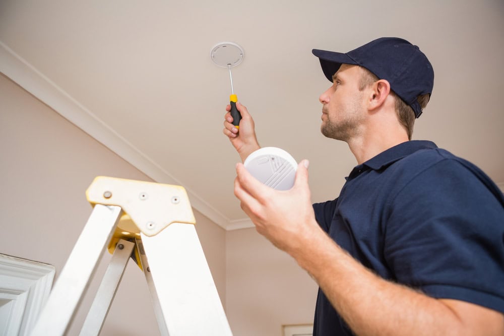 A man in a navy cap and shirt stands on a ladder installing a smoke detector on the ceiling with a screwdriver. He holds the smoke detector in one hand and works on the mounting with the other.