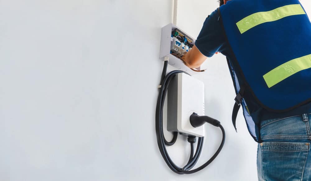 A technician wearing a blue vest installs a wall-mounted electric vehicle charging station. The charging cable is coiled and plugged in, with the technician adjusting wiring in a panel on the wall.