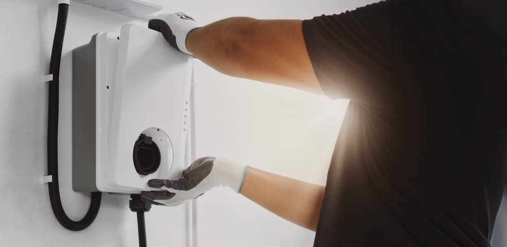 A person wearing gloves installs an electric vehicle charging station on a white wall. Sunlight highlights the scene from the background.