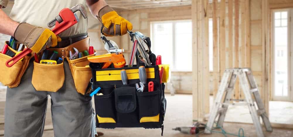 A person wearing a tool belt holds a toolbag filled with various tools in a partially constructed wooden room. A ladder stands in the background near large windows.