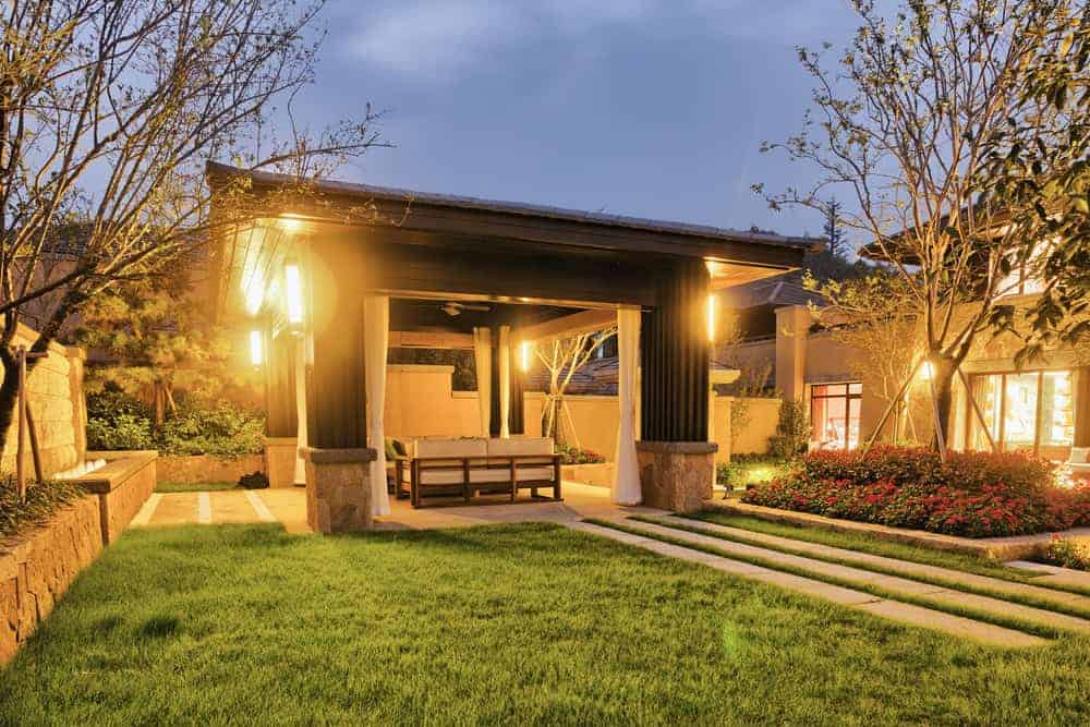 A well-lit backyard features a cozy wooden gazebo with a bench, surrounded by lush green grass and trees. Pathways lead to the gazebo, creating a serene setting. Plants and flowers are visible along the borders, and a house is seen in the background.