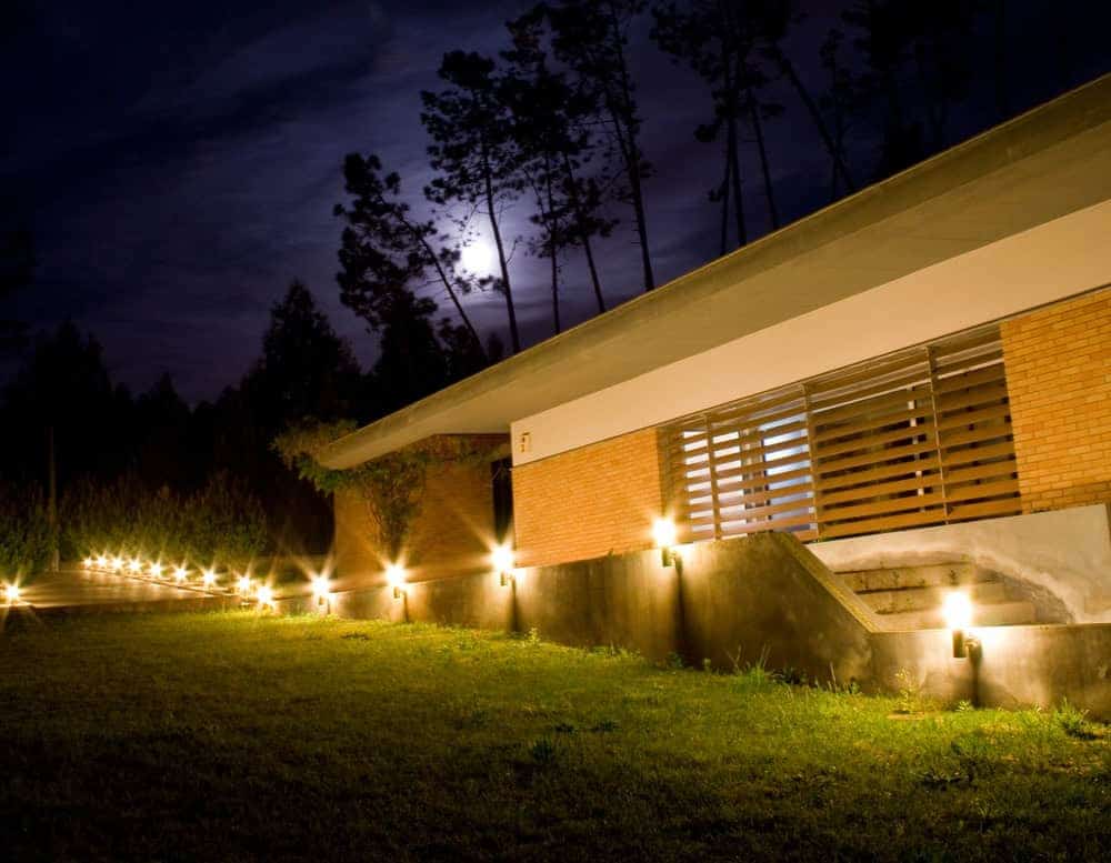 A modern house with a brick facade is lit by outdoor lights at night. The pathway is illuminated, and the moon is visible in the sky above silhouetted trees.