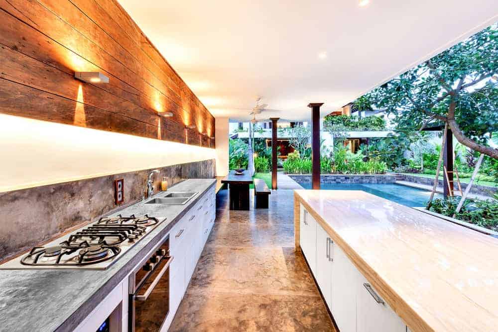 A modern open-air kitchen with stainless steel appliances and a large island with a marble countertop. It opens to a garden with a spacious pool surrounded by lush greenery and contemporary outdoor furniture.