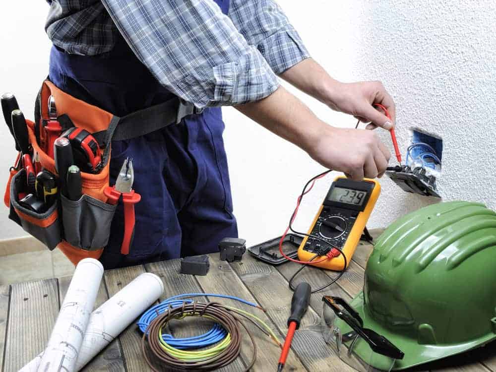 An electrician is working on a wall socket, using a multimeter to measure voltage. Nearby are a green hard hat, electrical wires, tools, and construction plans on a wooden table. The electrician wears a tool belt with various tools.